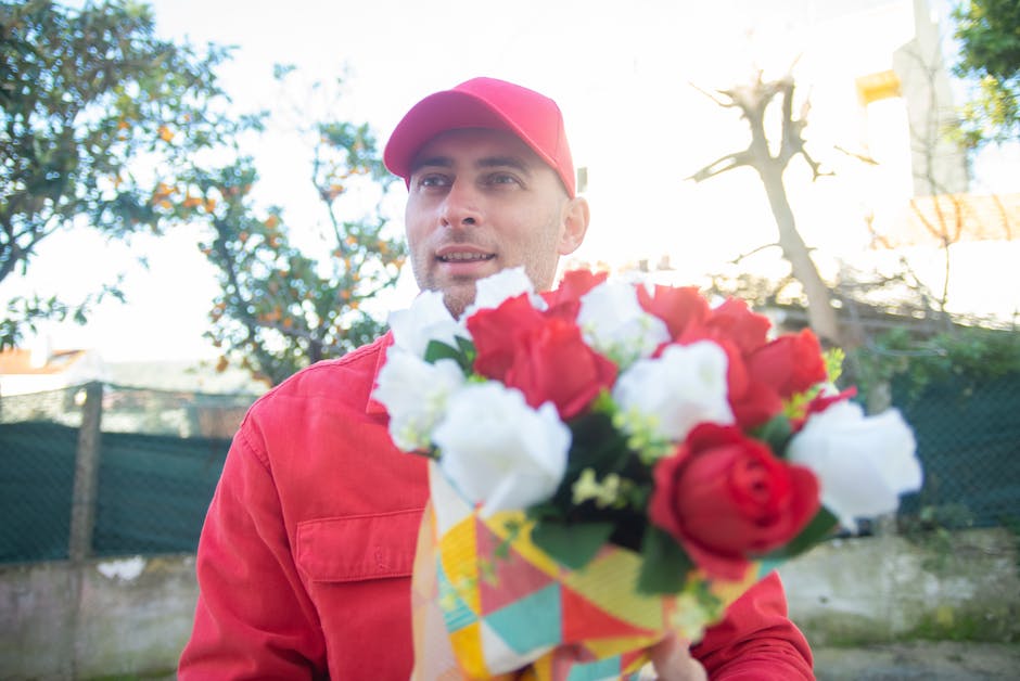 A delivery person dressed in a red uniform and cap is holding a vibrant floral bouquet consisting of red and white roses with green foliage, wrapped in colorful patterned paper. The bouquet is positio