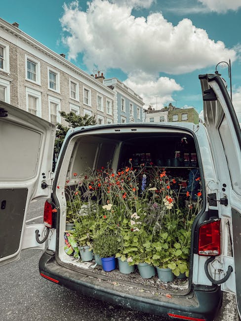 The open rear of a white delivery van filled with potted flowers, including tall stems of red and white poppies and other green foliage plants, arranged in light blue and white pots, set against the b