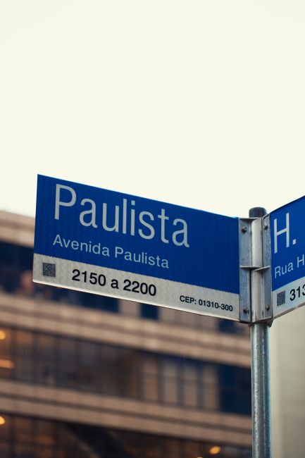 A close-up of a blue street sign reading 'Paulista Avenida Paulista', mounted on a metal pole against a background of modern buildings with glass facades. The sign indicates the street's location and 
