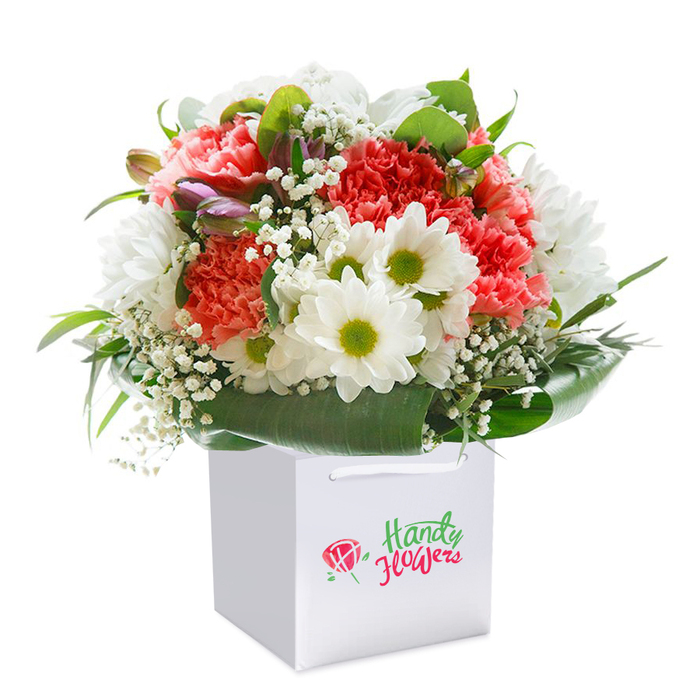 A captivating floral display emerges from a pristine white square gift bag, effortlessly blending the lively energy of South Tottenham with the tranquil artistry of expert floristry. The arrangement overflows with peach and coral carnations, their soft, frilled petals lending texture and warmth that recall the sunlit gardens of Downhills Park. Bright white daisies with vivid lime and yellow-green centers burst among the carnations, bringing playful contrast and a mood of fresh optimism-just as you might feel wandering the side streets between Turnpike Lane and Tottenham Green. Airy sprigs of baby's breath appear throughout the bouquet like delicate wisps of morning mist, offering an understated lightness that elevates the whole creation. Subtle, tightly closed buds brushed with hints of purple and rose add dimension and a quiet sense of unfolding possibility. Deep, shiny green leaves encircle the entire arrangement, providing a rich, botanical marrow that frames the flowers and ties the presentation together with poise. Every element is professionally arranged in a sturdy white gift bag with smooth handles, suggesting both elegance and ease of delivery-ready to be gifted for a birthday, anniversary, or thoughtful gesture anywhere across South Tottenham. The branding is discrete, allowing the vivid colours, masterful textures, and sense of occasion to shine through. This bouquet is a testament to the freshness and creativity that define local floral delivery, offering recipients more than flowers: it's a piece of South Tottenham's blooming, heartfelt spirit.
