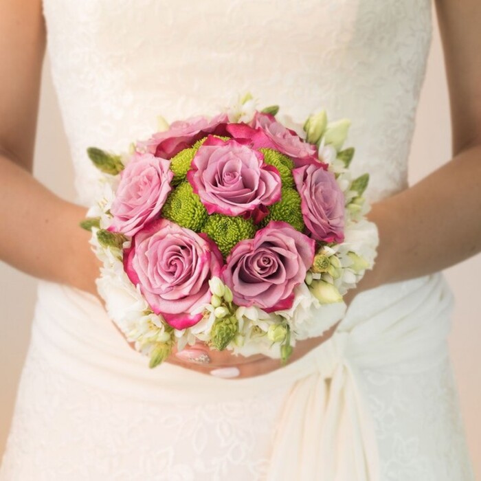 A waist-level view in soft, natural light reveals a bride in South Tottenham holding her wedding bouquet before a backdrop of elaborate ivory lace-a sight often admired at celebrations near Bruce Castle Park. The bouquet is composed with a flair for colour harmony and texture, centering on full-bloomed roses shaded from gentle lavender to rosy mauve and crowned with vivid fuchsia at the edges. These luxurious blooms exude romance, surrounded by a lively ring of lime-green button chrysanthemums, their dense, compact forms providing an energetic pop and tactile contrast that feels as fresh as a spring market morning on High Road. Encircling this vibrant core, a halo of petite white flowers-freesia, waxflower, or small spray roses-provides a gossamer finish, enhanced by slender green buds and leaves that recall the wild beauty of local gardens. The bride's hands are gracefully poised, nails painted a tender pink with a hint of sparkle-each gesture resonating with anticipation and joy. The gentle drape of her dress and the interplay of lace detail elevate the scene's elegance and intimacy. This moment combines classic bridal tradition with the thoughtful selection found in South Tottenham's skilled florists-a homage to local artistry that transforms natural blooms into symbols of everlasting love and celebration.